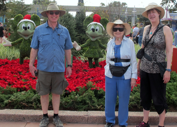 Picture of Peter Burck, his mother, Dorothy, and his wife, Susan Hoines in front of topiaries of Mickey Mouse and Minnie Mouse at Epcot. Behind them is the Christmas tree, cut off at the top. The Monorail appears behind them at the level of their heads.